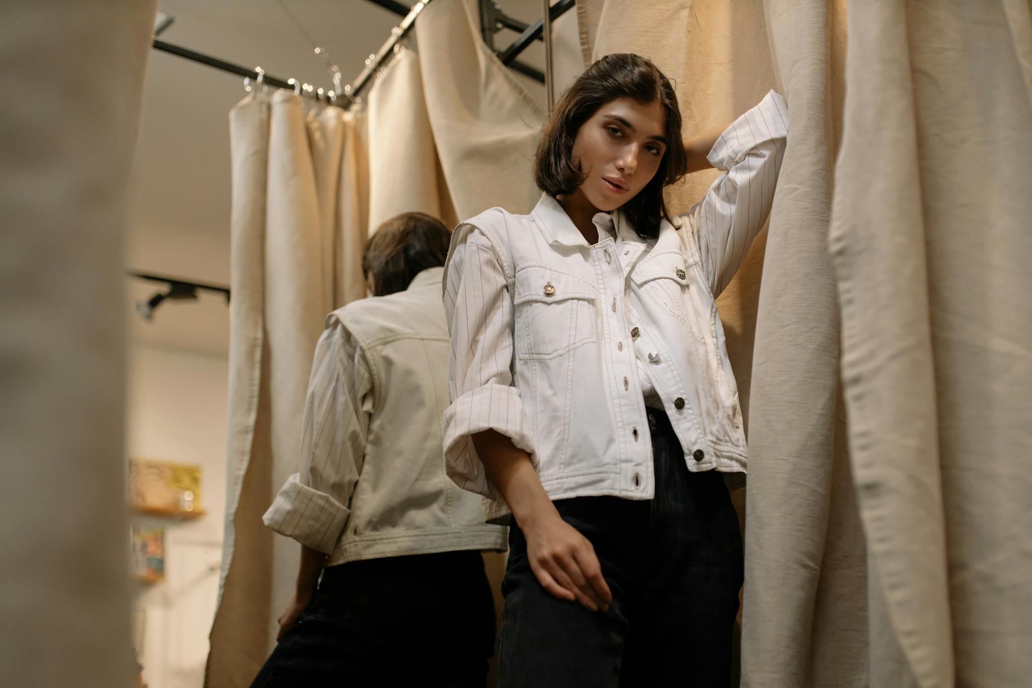 A stylish woman in a casual outfit poses inside a fitting room with curtains in a clothing store.