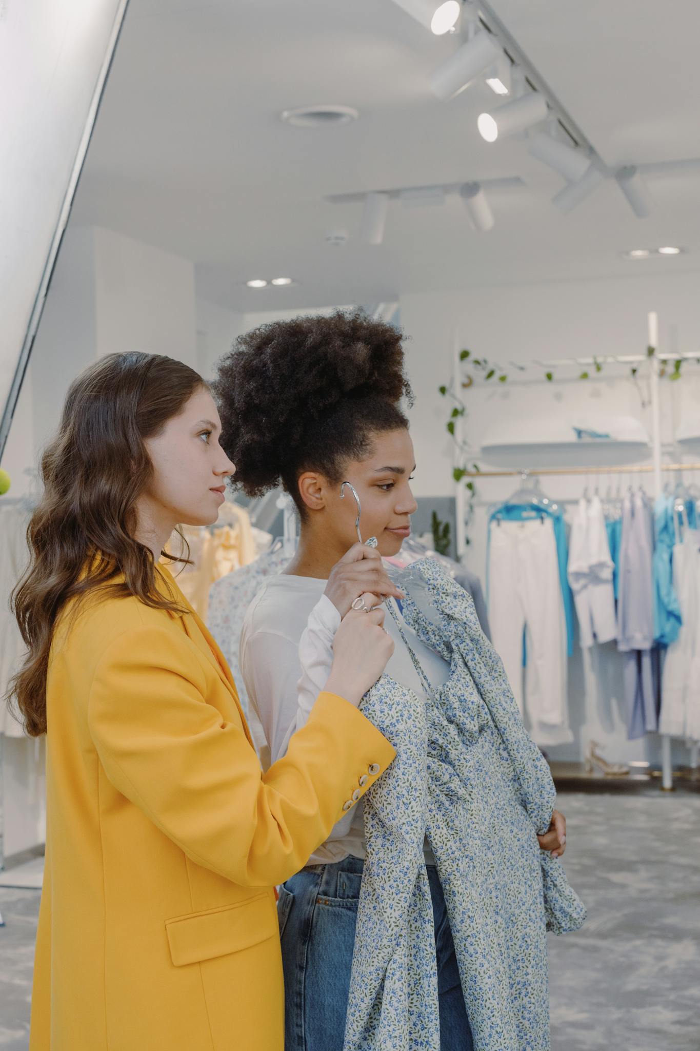 Two women shopping in a clothing store, picking out outfits and styling each other with friendly interaction.